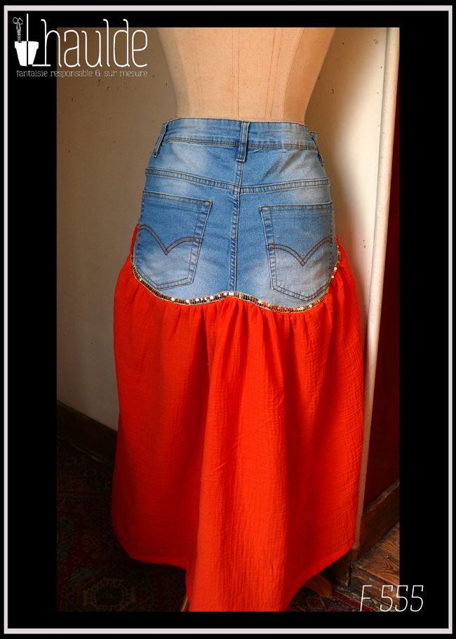 Jupe mi-longue, vue de dos, le haut est un jean coupé juste au-dessus de l'entrejambe, les courbes des fesses sont conservées. Le bas est en double gaze orange, un peu plus long que devant et non plissé Un galon de sequin doré marque la séparation entre les deux tissus Vue de dos sur un mannequin de couture