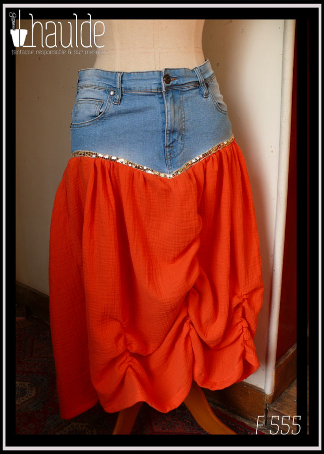 Jupe mi-longue, le haut est un jean coupé juste au-dessus de l'entrejambe avec des courbes qui remontent vers les côtés, le bas est en double gaze orange plissée à la verticale, trois fois, au centre et de chaque côté. Un galon de sequin doré marque la séparation entre les deux tissus Vue de face sur un mannequin de couture