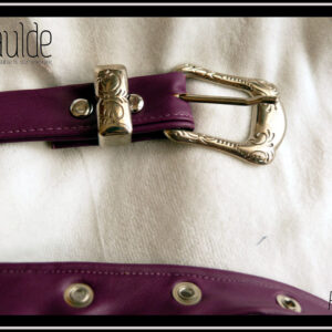 Vue de détail de la boucle de la ceinture en simili cuir violet, boucle rectangulaire en acier martelé (motifs arabesques végétales) Des oeillets d'acier protège les trous. Vue posée sur du velours blanc