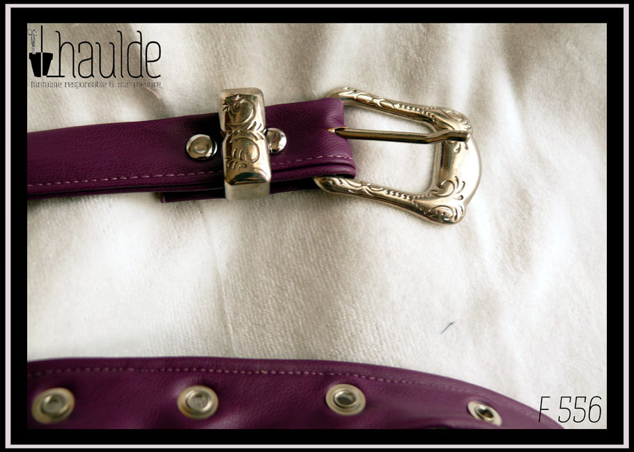 Vue de détail de la boucle de la ceinture en simili cuir violet, boucle rectangulaire en acier martelé (motifs arabesques végétales) Des oeillets d'acier protège les trous. Vue posée sur du velours blanc