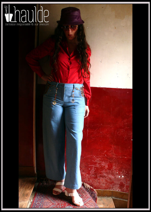 Jean bleu clair avec une ceinture bijou dont les extrémités pendant sur l'avant du jean Chemisier de satin de coton rouge cerise, bob en simili cuir violet Le tout porté par une femme