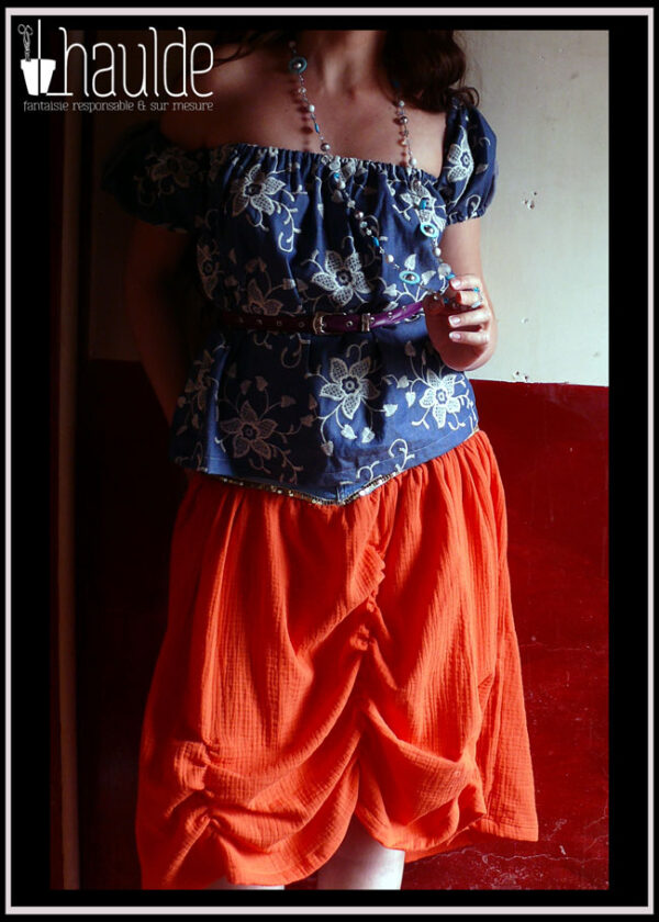 Haut en jean brodé de fleurs de dentelle blanche, décolleté élastique, manches bouffantes qui peuvent se rabattre sur les bras Jupe en double gaze orange plissée à la verticale, ceinture fine en simili cuir violet, long collier de perles de verre, métal et nacre Vus portés par une femme dont on ne voit ni la tête ni les jambes