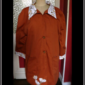 Manteau en sergé roux, col et manchettes blanches imprimé ginkgo, le même imprimé est découpé et appliqué en bas du pan avant droit Deux grandes poches plaquées, deux boutons Vu de face sur un mannequin de couture