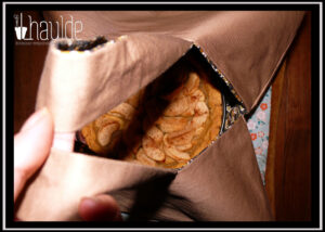 Sac à tarte en sergé beige, intérieur imprimé petites fleurs ocres sur fond noir, deux anses Vu tenu en l'air par les anses, à travers la fente on aperçoit une tarte aux pommes