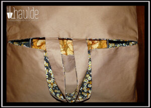 Sac à tarte en sergé beige, intérieur imprimé petites fleurs ocres sur fond noir, deux anses, à travers la fente on aperçoit une tarte aux pommes. Vu de dessus