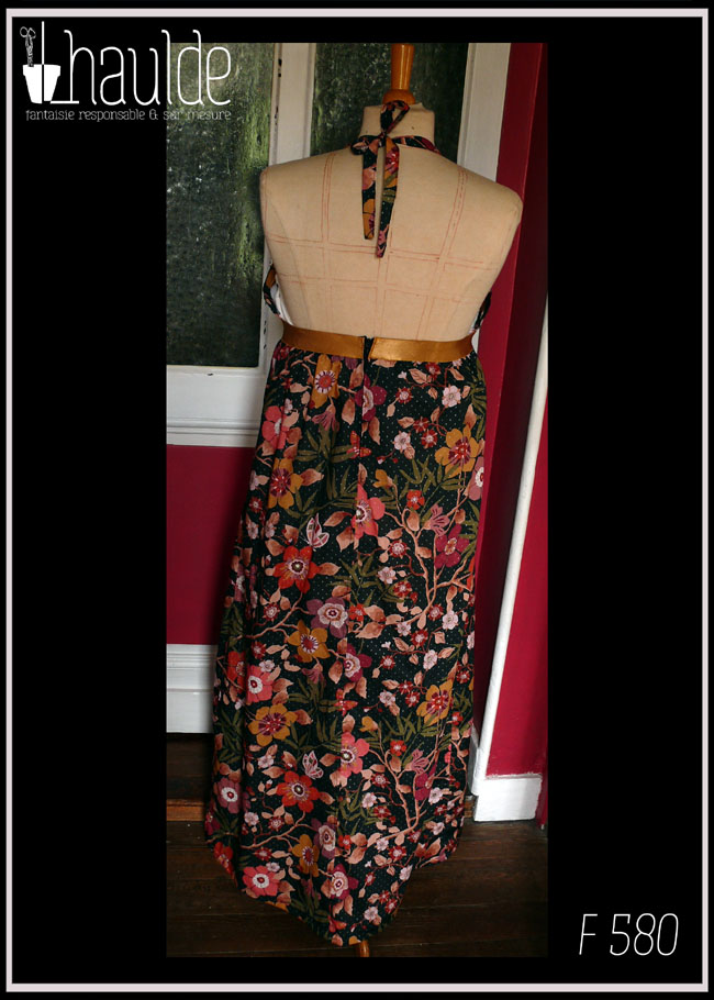 Robe longue dos nu, bretelles nouées au cou en tissu Liberty™ noir imprimé motifs fleuris et petits pois dorés Une bande élastique dorée ceint la robe sous la poitrine Vue de face sur un mannequin de couture