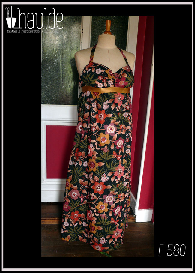 Robe longue dos nu, bretelles nouées au cou en tissu Liberty™ noir imprimé motifs fleuris et petits pois dorés Une bande élastique dorée ceint la robe sous la poitrine Vue de face sur un mannequin de couture