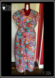 Robe longue manches très courtes en tissu imprimé fleurs stylisées multicolores (dominante rose et orange-rouge) et feuillage turquoise sur fond rose. Vue de face sur un mannequin de couture Une ceinture dans le même tissu, boucle ronde en plastique bleu marine complète la tenue. Encolure en V arrondi