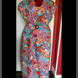 Robe longue manches très courtes en tissu imprimé fleurs stylisées multicolores (dominante rose et orange-rouge) et feuillage turquoise sur fond rose. Vue de face sur un mannequin de couture Une ceinture dans le même tissu, boucle ronde en plastique bleu marine complète la tenue. Encolure en V arrondi