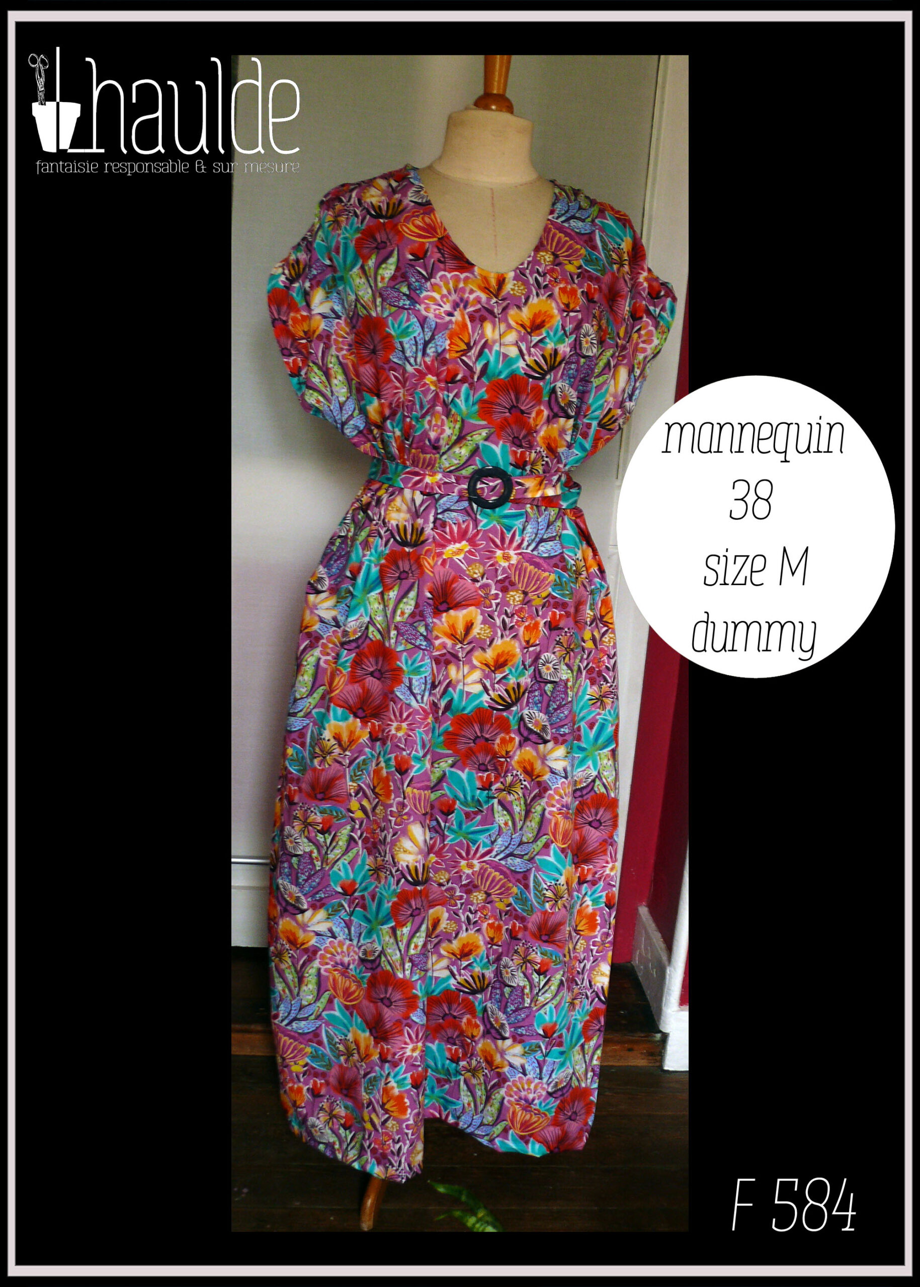 Robe longue manches très courtes en tissu imprimé fleurs stylisées multicolores (dominante rose et orange-rouge) et feuillage turquoise sur fond rose. Vue de face sur un mannequin de couture qui est cinq tailles en dessous de la robe (un 38 au lieu d'un 48) pour montrer qu'elle peut aussi se porter comme ça Une ceinture dans le même tissu, boucle ronde en plastique bleu marine complète la tenue et permet de l'ajuster Encolure en V arrondi qui sur une petite taille parait franchement rond