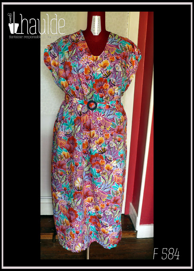 Robe longue manches très courtes en tissu imprimé fleurs stylisées multicolores (dominante rose et orange-rouge) et feuillage turquoise sur fond rose. Vue de face sur un mannequin de couture Une ceinture dans le même tissu, boucle ronde en plastique bleu marine complète la tenue. Encolure en V arrondi