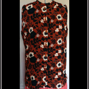 Haut sans manches, froncé aux épaules en viscose terracotta imprimée fleurs blanches et silhouettes de fleurs et feuilles noires Boutons en résine contenant des fleurs séchées oranges. Col à liseret simple Vu de face sur un mannequin de couture