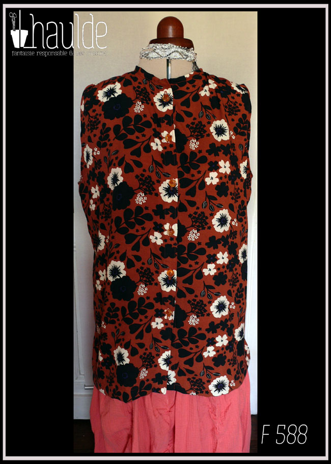 Haut sans manches, froncé aux épaules en viscose terracotta imprimée fleurs blanches et silhouettes de fleurs et feuilles noires Boutons en résine contenant des fleurs séchées oranges. Col à liseret simple Vu de face sur un mannequin de couture