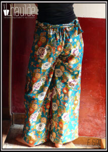 Pantalon ample en coton vert canard imprimé de camélia japonais blancs, vermillon, or et bleus cernés d'or Il est noué des deux côté à la taille Vu porté, de dos, la taille est élastiquée dans le dos ce qui crée des plis au niveau des reins