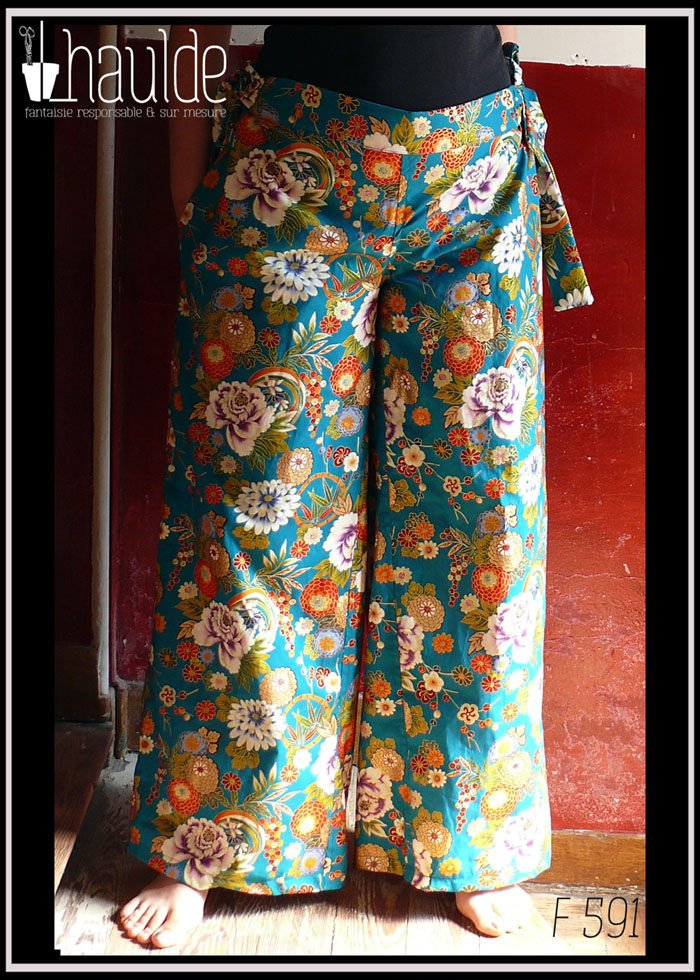 Pantalon ample en coton vert canard imprimé de camélia japonais blancs, vermillon, or et bleus cernés d'or Il est noué des deux côté à la taille, ici avec les pans de la ceinture qui pendent Vu porté, de face