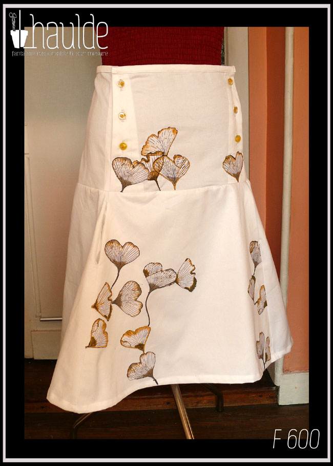 Jupe mi-longue en sergé blanc, ajustée jusqu'aux hanches puis plus ample, avec sur le devant des appliqués de tissu imprimé feuilles de ginkgo translucides. De la taille aux hanches, de chaque côté, trois rangées de boutons en résine à l'intérieur desquels sont incrustés de petites fleurs jaunes (boutons de chez Ortilys) Vue de trois quarts sur un mannequin de couture