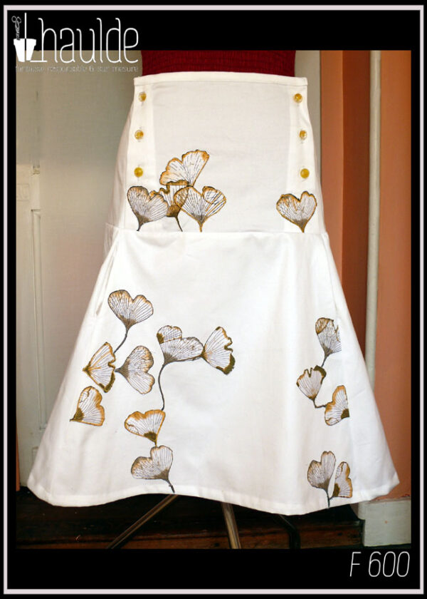 Jupe mi-longue en sergé blanc, ajustée jusqu'aux hanches puis plus ample, avec sur le devant des appliqués de tissu imprimé feuilles de ginkgo translucides. De la taille aux hanches, de chaque côté, trois rangées de boutons en résine à l'intérieur desquels sont incrustés de petites fleurs jaunes (boutons de chez Ortilys) Vue de face sur un mannequin de couture