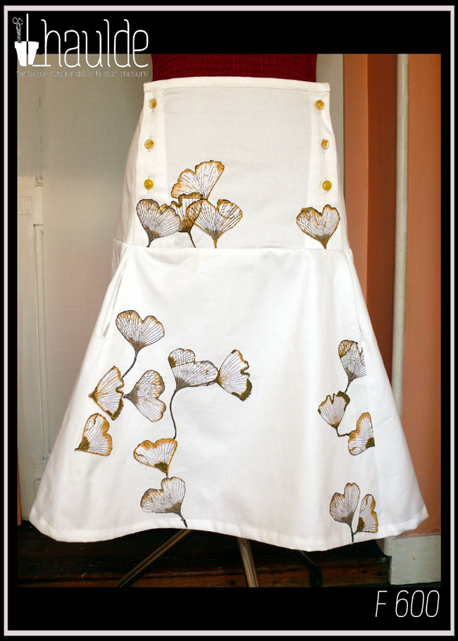 Jupe mi-longue en sergé blanc, ajustée jusqu'aux hanches puis plus ample, avec sur le devant des appliqués de tissu imprimé feuilles de ginkgo translucides. De la taille aux hanches, de chaque côté, trois rangées de boutons en résine à l'intérieur desquels sont incrustés de petites fleurs jaunes (boutons de chez Ortilys) Vue de face sur un mannequin de couture
