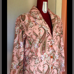 La veste vue de trois quarts face sur un mannequin de couture Manches longues, tissu sergé rose pâle à motifs végétaux et paisley dans les tons verts kakis et roses avec des fleurs blanches Trois boutons en résine avec des fleurs oranges incrustées dedans