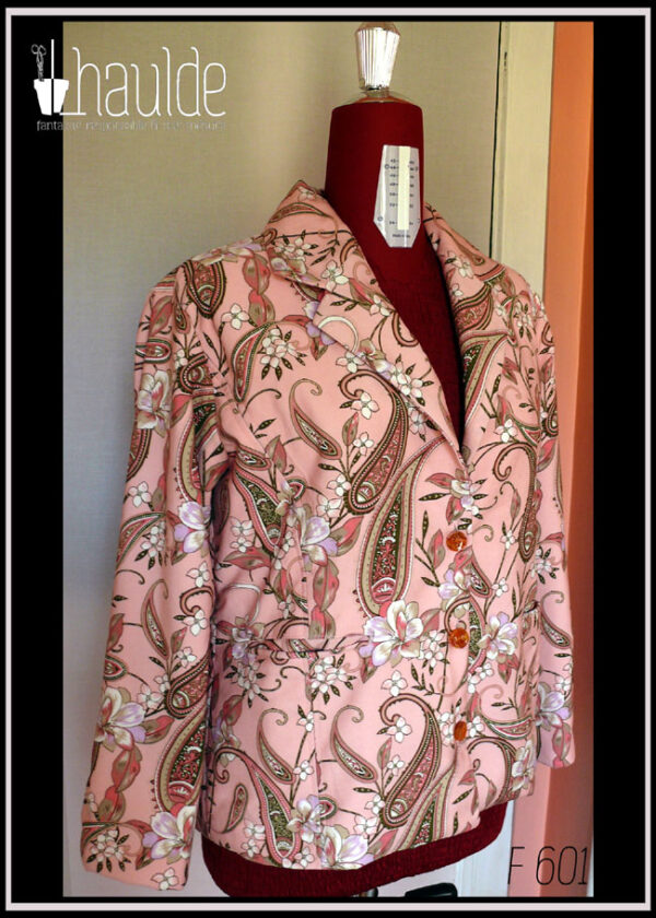 La veste vue de trois quarts face sur un mannequin de couture Manches longues, tissu sergé rose pâle à motifs végétaux et paisley dans les tons verts kakis et roses avec des fleurs blanches Trois boutons en résine avec des fleurs oranges incrustées dedans