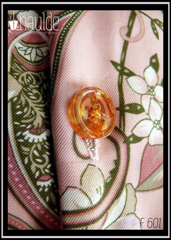 Gros plan d'un bouton en résine avec des pétales de fleurs oranges incrustées dedans boutonné sur un tissu sergé rose pâle à motifs végétaux et paisley dans les tons verts kakis et roses