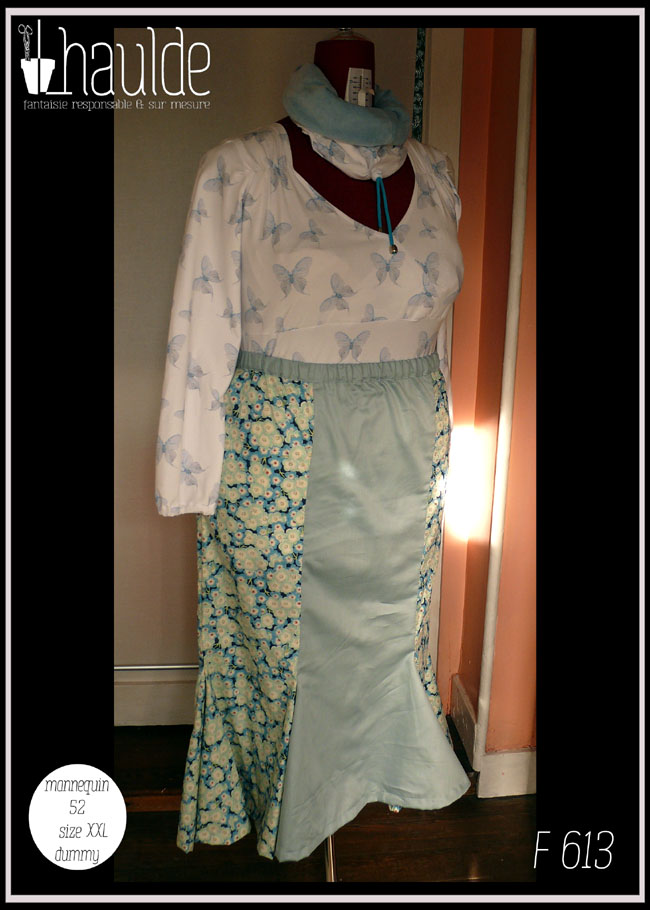 Jupe longue à taille élastique et à double quilles en bas en coton bleu imprimé petites fleurs de cerisiers blanches. Au centre devant il y a un pan de satin bleu pâle. La ceinture est dans la même matière Haut manches longues coeur croisé en jersey blanc imprimé papillons bleus translucides Il y a une légère fronce de chaque côté sous les épaules et les manchettes sont élastiquées Vu de trois quarts face sur un mannequin de couture réglé sur la taille 52. Le haut est très moulant et passe sous la jupe Un macaron en bas à gauche indique que la photo a été prise sur un mannequin de couture réglé en taille 52