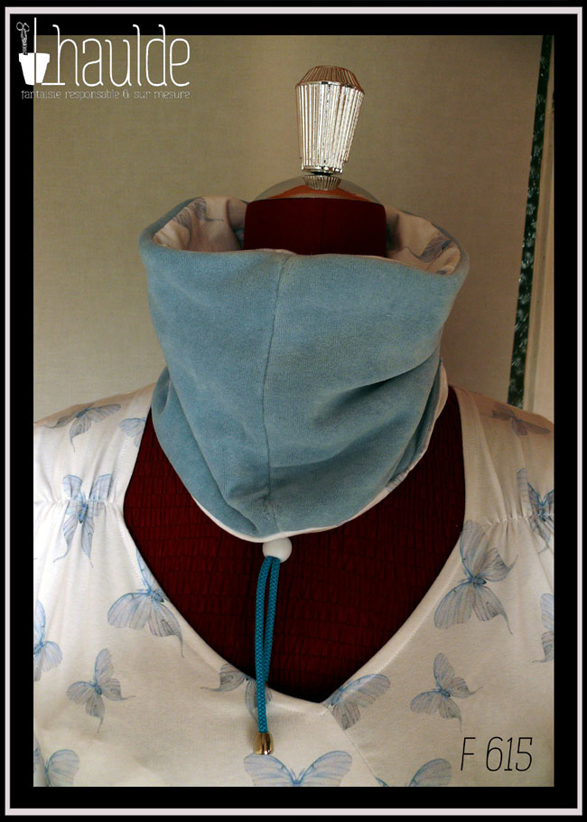 Tour de cou vu côté velours bleu pastel. On aperçoit également le cordon de serrage bleu et le stop en plastique blanc Le cache col est posé sur un mannequin de couture qui porte un haut assorti en jersey blanc imprimé papillons bleus (référence F 614)