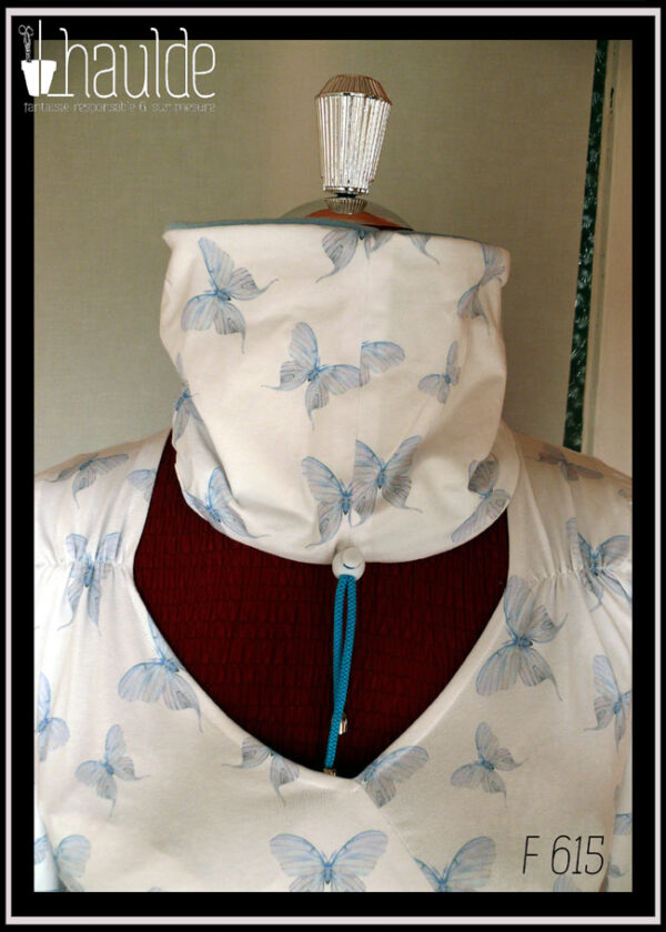 Tour de cou vu relevé côté jersey blanc imprimé papillons bleus. On aperçoit également le cordon de serrage bleu et le stop en plastique blanc Le cache col est posé sur un mannequin de couture qui porte un haut assorti en jersey blanc imprimé papillons bleus (référence F 614)