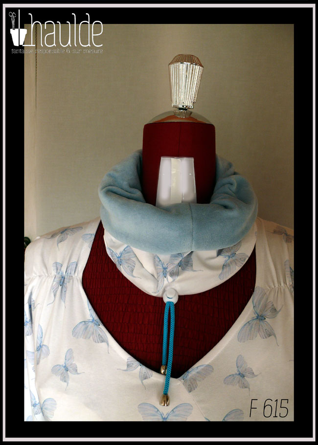 Tour de cou vu replié, le velours bleu pastel sur le jersey imprimé. On aperçoit également le cordon de serrage bleu et le stop en plastique blanc Le cache col est posé sur un mannequin de couture qui porte un haut assorti en jersey blanc imprimé papillons bleus (référence F 614)