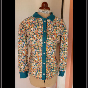 Chemisier à manches longues en tissu bleu paon imprimé fleurs stylisées dans des cercles, d'inspiration japonaise. Elles sont multicolores avec des dominantes oranges et blanches. La boutonnière, les manchettes et le col sont en tissu uni bleu paon. Les boutons sont en résine transparente rehaussée de blanc et remplis de fleurs séchées multicolores. Ils viennent de chez Ortilys Vue de face sur un mannequin de couture réglé en taille 36