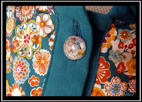 Vue de détail d'un autre bouton Chemisier à manches longues en tissu bleu paon imprimé fleurs stylisées dans des cercles, d'inspiration japonaise. Elles sont multicolores avec des dominantes oranges et blanches. La boutonnière, les manchettes et le col sont en tissu uni bleu paon. Les boutons sont en résine transparente rehaussée de blanc et remplis de fleurs séchées multicolores. Ils viennent de chez Ortilys