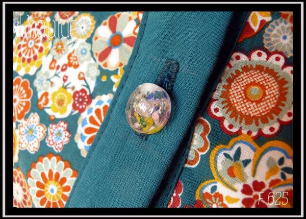 Vue de détail d'un des boutons Chemisier à manches longues en tissu bleu paon imprimé fleurs stylisées dans des cercles, d'inspiration japonaise. Elles sont multicolores avec des dominantes oranges et blanches. La boutonnière, les manchettes et le col sont en tissu uni bleu paon. Les boutons sont en résine transparente rehaussée de blanc et remplis de fleurs séchées multicolores. Ils viennent de chez Ortilys