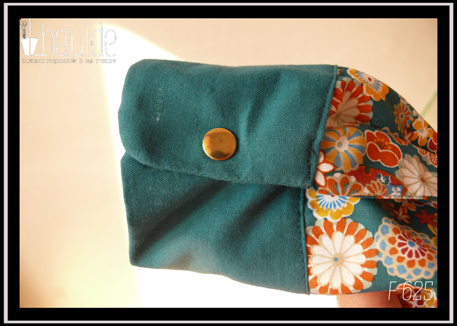 Détail de la manchette, fermée par un bouton pression doré Chemisier à manches longues en tissu bleu paon imprimé fleurs stylisées dans des cercles, d'inspiration japonaise. Elles sont multicolores avec des dominantes oranges et blanches. La boutonnière, les manchettes et le col sont en tissu uni bleu paon. Les boutons sont en résine transparente rehaussée de blanc et remplis de fleurs séchées multicolores. Ils viennent de chez Ortilys