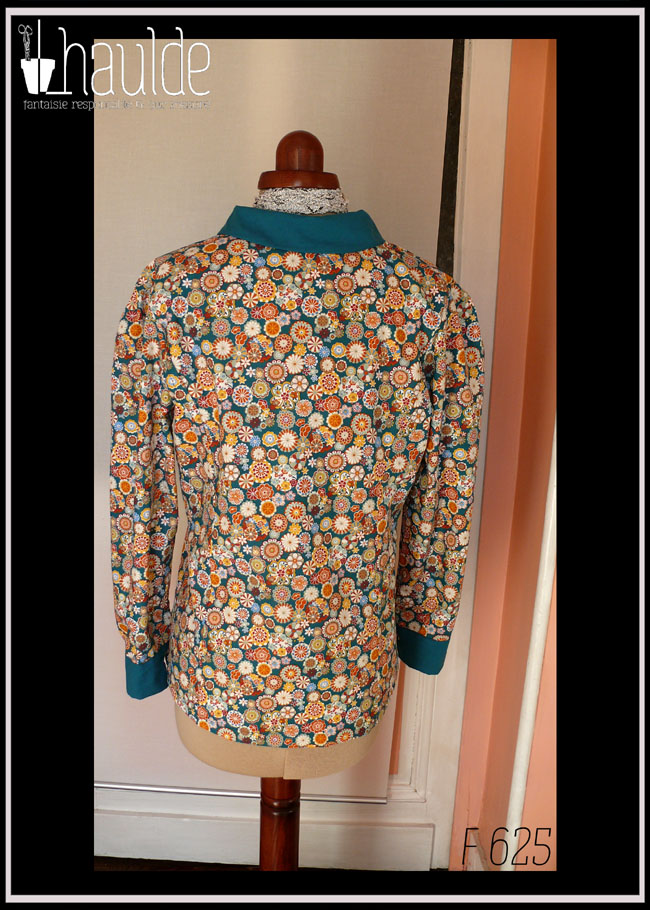 Chemisier à manches longues en tissu bleu paon imprimé fleurs stylisées dans des cercles, d'inspiration japonaise. Elles sont multicolores avec des dominantes oranges et blanches. La boutonnière, les manchettes et le col sont en tissu uni bleu paon. Les boutons sont en résine transparente rehaussée de blanc et remplis de fleurs séchées multicolores. Ils viennent de chez Ortilys Vue de dos sur un mannequin de couture réglé en taille 36