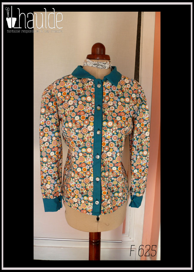 Chemisier à manches longues en tissu bleu paon imprimé fleurs stylisées dans des cercles, d'inspiration japonaise. Elles sont multicolores avec des dominantes oranges et blanches. La boutonnière, les manchettes et le col sont en tissu uni bleu paon. Les boutons sont en résine transparente rehaussée de blanc et remplis de fleurs séchées multicolores. Ils viennent de chez Ortilys Vue de face sur un mannequin de couture réglé en taille 36