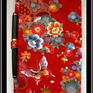 Protège carnet en tissu rouge imprimé fleurs et papillons d'esthétique japonaise. Vu fermé avec un stylo dans l'anneau situé sur le bord du rabat