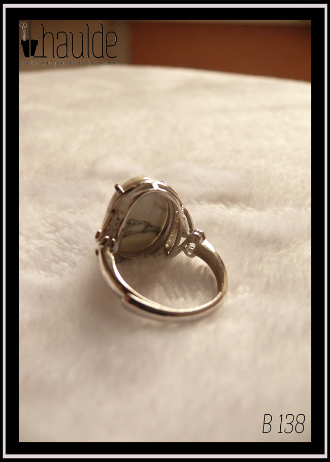 Bague avec un cabochon ovale en pierre naturelle blanche marbrée de gris (de la howlite) montée sur un support en argent bordé de petits éclats d'oxyde de zirconium. Des arabesques font filer le dessus de la bague vers l'anneau réglable Vue de dos, on voit que l'anneau est réglable