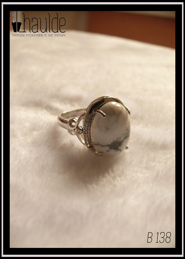 Bague avec un cabochon ovale en pierre naturelle blanche marbrée de gris (de la howlite) montée sur un support en argent bordé de petits éclats d'oxyde de zirconium. Des arabesques font filer le dessus de la bague vers l'anneau réglable Vue sur fond blanc La pierre est opaque