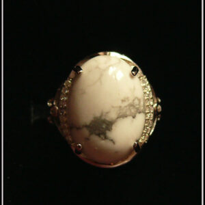 Bague avec un cabochon ovale en pierre naturelle blanche marbrée de gris (de la howlite) montée sur un support en argent bordé de petits éclats d'oxyde de zirconium. Des arabesques font filer le dessus de la bague vers l'anneau réglable Vue sur fond noir