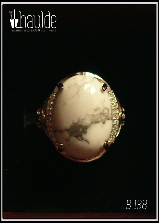 Bague avec un cabochon ovale en pierre naturelle blanche marbrée de gris (de la howlite) montée sur un support en argent bordé de petits éclats d'oxyde de zirconium. Des arabesques font filer le dessus de la bague vers l'anneau réglable Vue sur fond noir
