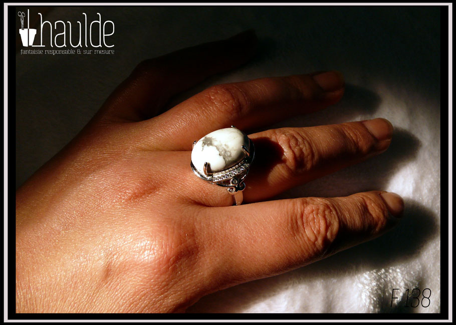 Bague avec un cabochon ovale en pierre naturelle blanche marbrée de gris (de la howlite) montée sur un support en argent bordé de petits éclats d'oxyde de zirconium. Des arabesques font filer le dessus de la bague vers l'anneau réglable Vue portée sur un majeur, au soleil