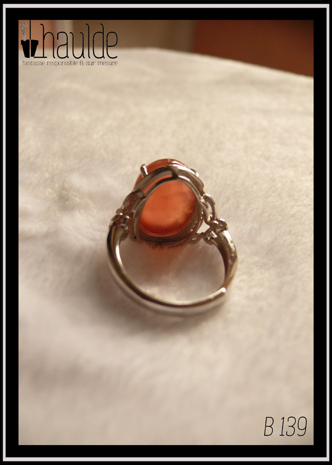 Bague avec un cabochon ovale en pierre naturelle rose-rouge translucide (de la rhodochrosite) montée sur un support en argent bordé de petits éclats d'oxyde de zirconium. Des arabesques font filer le dessus de la bague vers l'anneau réglable Bague vue de dos, la pierre est translucide, on voit que l'anneau peut se régler