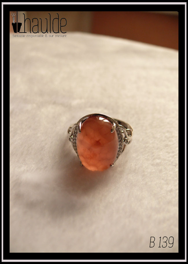 Bague avec un cabochon ovale en pierre naturelle rose-rouge translucide (de la rhodochrosite) montée sur un support en argent bordé de petits éclats d'oxyde de zirconium. Des arabesques font filer le dessus de la bague vers l'anneau réglable Vue sur un fond blanc, la translucidité de la pierre est très visible