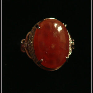Bague avec un cabochon ovale en pierre naturelle rose-rouge translucide (de la rhodochrosite) montée sur un support en argent bordé de petits éclats d'oxyde de zirconium. Des arabesques font filer le dessus de la bague vers l'anneau réglable Vue sur fond noir