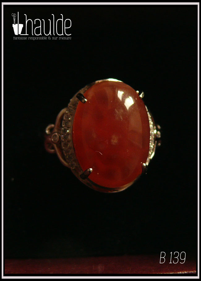 Bague avec un cabochon ovale en pierre naturelle rose-rouge translucide (de la rhodochrosite) montée sur un support en argent bordé de petits éclats d'oxyde de zirconium. Des arabesques font filer le dessus de la bague vers l'anneau réglable Vue sur fond noir