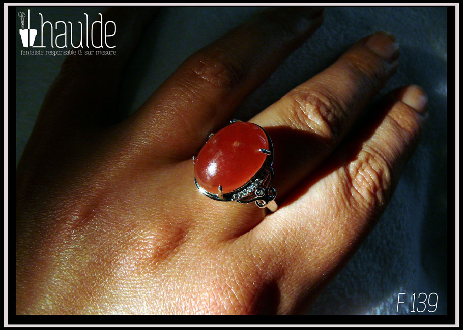 Bague avec un cabochon ovale en pierre naturelle rose-rouge translucide (de la rhodochrosite) montée sur un support en argent bordé de petits éclats d'oxyde de zirconium. Des arabesques font filer le dessus de la bague vers l'anneau réglable Vue portée, sur un majeur, au soleil
