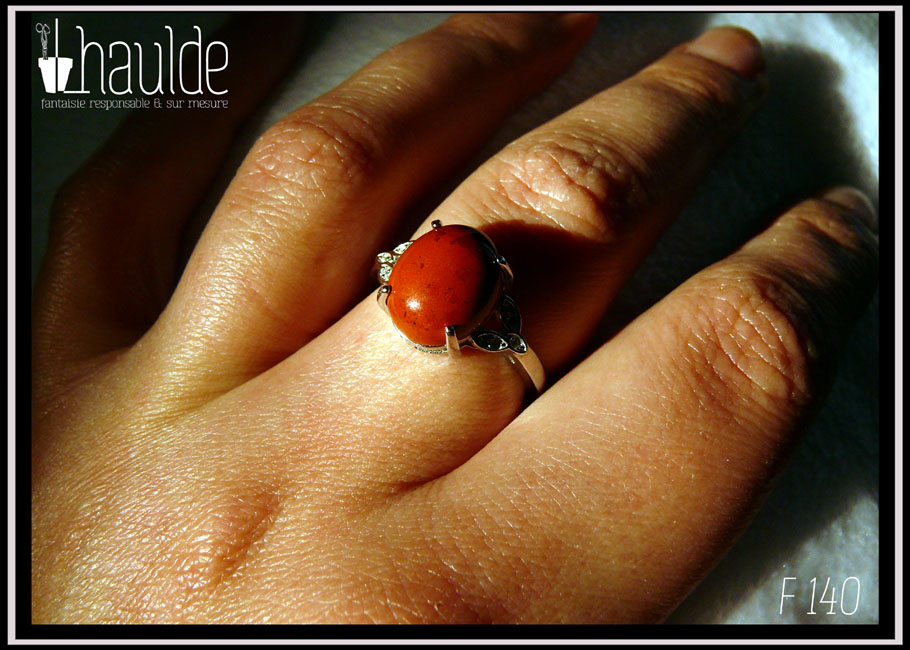Bague avec un petit cabochon ovale en jaspe rouge monté sur un support en argent garni de chaque côté de trois petites mandorles contenant des éclats d'oxyde de zirconium vue portée sur un majeur au soleil
