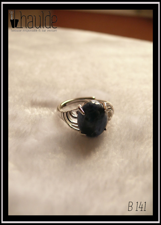 Bague avec un petit cabochon ovale en sodalite, pierre bleu foncé marbré de bleu nuit, montée sur un support en argent dont les côtés sont ajourés. Vue de trois quarts face sur un support blanc, la pierre est opaque