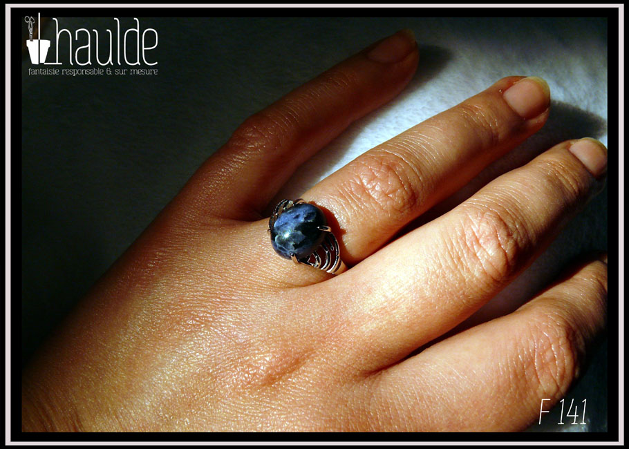 Bague avec un petit cabochon ovale en sodalite, pierre bleu foncé marbré de bleu nuit, montée sur un support en argent dont les côtés sont ajourés. Vue portée sur un annulaire au soleil
