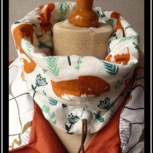 Tour de cou en tissu blanc imprimé renard roux et végétaux clairsemés dans les tons verts, le dessin est de style naïf, les renards sont dans plusieurs postures, devant il apparait couché, mais il y en a aussi des assis Cordon de serrage marron avec un stop blanc Vu sur un mannequin de couture